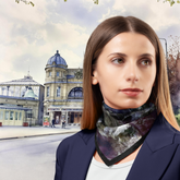 Woman wearing a scarf with a scenic print, standing in front of a vintage-style building.